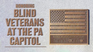 An American flag in bronze with the Pledge of Allegiance written in Braille, with the words “Honoring Blind Veterans At The PA Capitol” in bronze against a gray background.